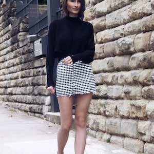 Pu&ograve; includere: Una donna indossa un maglione nero a collo alto, una gonna a quadri bianchi e neri e sandali di pelliccia neri. Sta camminando su un marciapiede di fronte a un muro di pietra.
