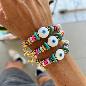 Può includere: Tre braccialetti colorati con ciondoli di occhio turco bianco e dettagli a stella marina dorati. I braccialetti hanno perline turchesi, rosa e bianche con motivi floreali.