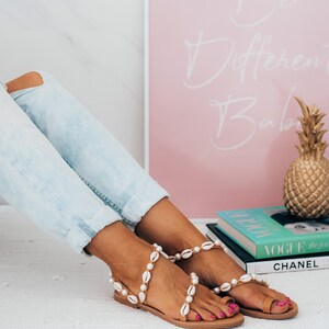 May include: A pair of tan sandals with white shell embellishments. The sandals are worn by a person wearing light blue jeans. The sandals are on a white surface with a pink framed poster that says "Be Different Babe" and a stack of books with the word "VOGUE" visible.