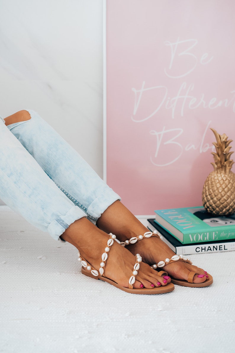 May include: A pair of tan sandals with white shell embellishments. The sandals are worn by a person wearing light blue jeans. The sandals are on a white surface with a pink framed poster that says "Be Different Babe" and a stack of books with the word "VOGUE" visible.