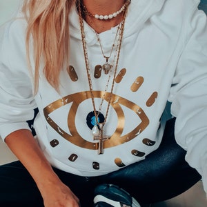 May include: A white hoodie with a gold eye graphic and a layered gold chain necklace with a pearl necklace and a small diamond pendant.