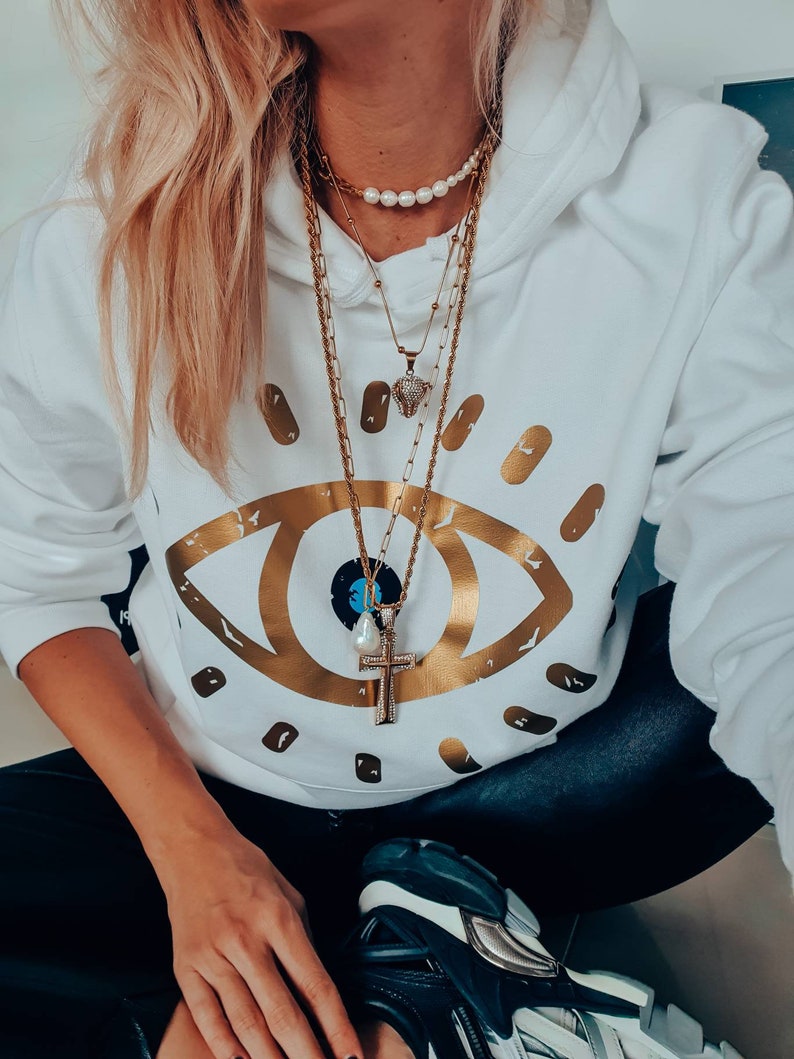 May include: A white hoodie with a gold eye graphic and a layered gold chain necklace with a pearl necklace and a small diamond pendant.