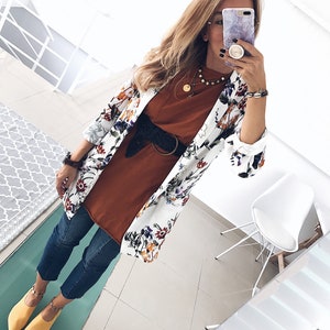 May include: A white floral print blazer with a brown t-shirt, blue jeans, and yellow shoes. The person is wearing a gold necklace and a black belt with a silver buckle.