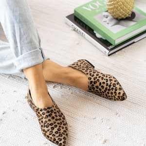 May include: A pair of brown and black leopard print flats with pointed toes. The shoes are on a white rug.