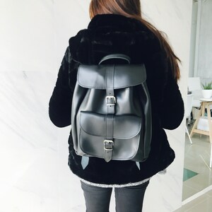 May include: A black leather backpack with two adjustable buckles and a flap closure. The backpack has a single pocket on the front.