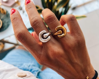 May include: Two abstract rings, one silver and one gold, worn on a finger. The rings are both open and have a curved shape.