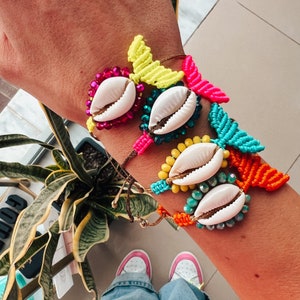 May include: A set of four colorful bracelets featuring seashells and macrame. The bracelets are made with bright pink, yellow, blue, and orange threads. Each bracelet has a different seashell charm.