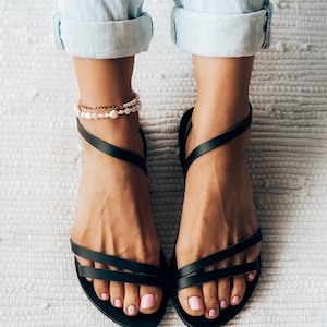 Puede incluir: Un par de sandalias de cuero negro con m&uacute;ltiples correas. Las sandalias son usadas por una persona con jeans de mezclilla remangados y tobilleras de perlas.