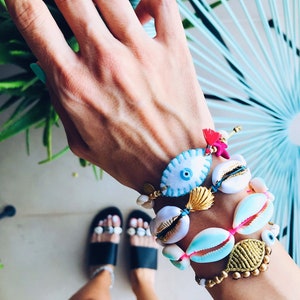 Op de afbeelding: Een hand met een stapel armbanden met blauwe en witte boze oogcharms, gouden schelpen en roze en gouden kralen.