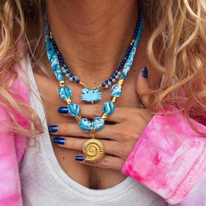 Puede incluir: Un collar de varias capas con cuentas azules y doradas. La capa superior presenta un dije de cangrejo azul, y la capa inferior tiene un dije de concha dorada.