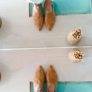May include: A pair of light blue jeans with distressed details are reflected in a mirror. The person wearing the jeans is standing on a tiled floor with two gold pineapple decorations. The person is wearing brown suede mules.