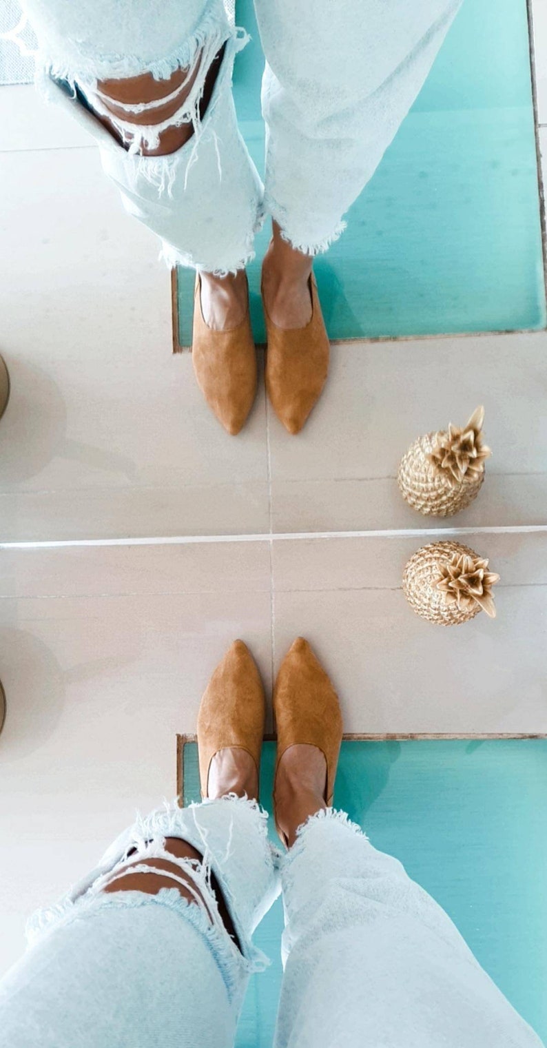 May include: A pair of light blue jeans with distressed details are reflected in a mirror. The person wearing the jeans is standing on a tiled floor with two gold pineapple decorations. The person is wearing brown suede mules.