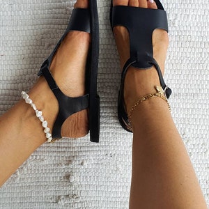 May include: A pair of black leather sandals with ankle straps and buckles. The sandals are on a white textured surface. One ankle has a pearl anklet and the other has a gold chain anklet.