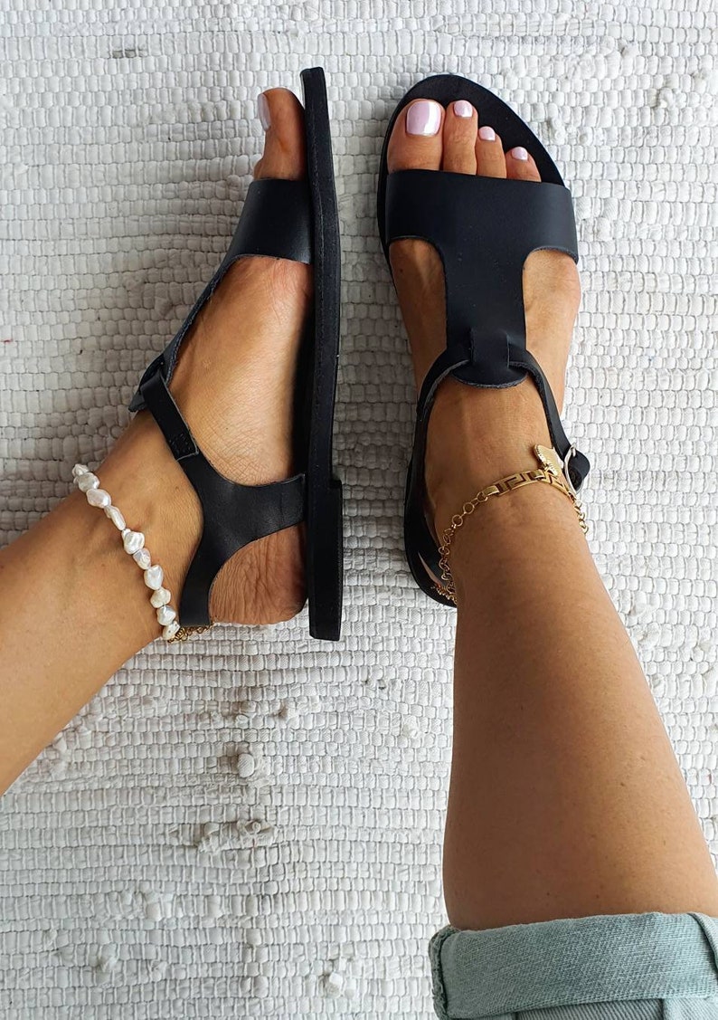 May include: A pair of black leather sandals with ankle straps and buckles. The sandals are on a white textured surface. One ankle has a pearl anklet and the other has a gold chain anklet.