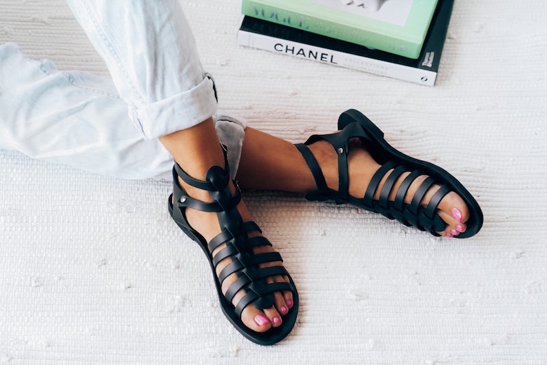 May include: A pair of black gladiator sandals with multiple straps. The sandals are made of a leather-like material and have a flat sole.