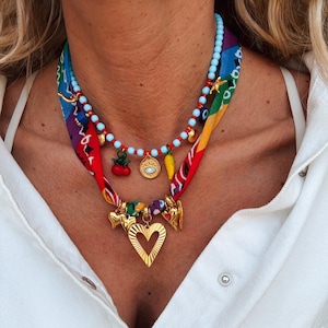 Può includere: Una collana a più strati con un design a sciarpa colorato. La collana presenta ciondoli dorati, tra cui un cuore, e vari ciondoli pendenti. La collana ha fili di perline blu e rosse. La sciarpa ha un motivo vibrante con tonalità rosse, blu, gialle e viola.