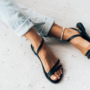 Puede incluir: Un par de sandalias de cuero negro con tiras finas. Las sandalias las lleva una persona con los pies descalzos y unos vaqueros azules claros.