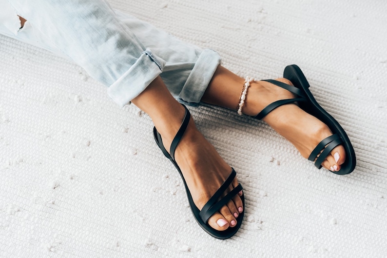 Puede incluir: Un par de sandalias de cuero negro con tiras finas. Las sandalias las lleva una persona con los pies descalzos y unos vaqueros azules claros.