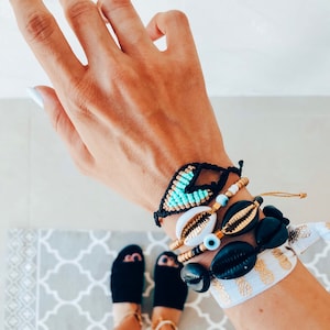 Op de afbeelding: Een hand draagt ​​een stapel armbanden met zwarte, witte en blauwe kralen, schelpen en een blauwe kwade oog-bedel. De armbanden zitten op een pols met een wit en gouden ananas-patroonband.
