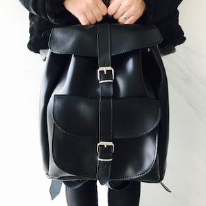 May include: A black leather backpack with silver buckles and a flap closure. The backpack has a front pocket with a flap and buckle.
