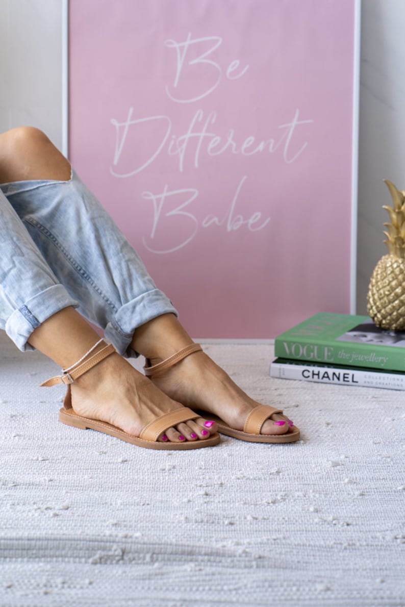 May include: A pair of tan leather sandals with ankle straps. The sandals are on a white rug with a pink framed poster in the background that says "Be Different Babe".