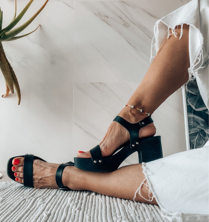 May include: A woman wearing black leather sandals with a chunky heel and a beaded ankle bracelet. The sandals have a strap across the toes and an ankle strap.