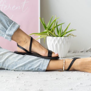 Puede incluir: Un par de sandalias de cuero negro con tiras finas. Las sandalias son usadas por una persona con los pies descalzos y un par de jeans azul claro.