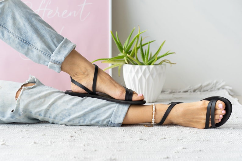 Puede incluir: Un par de sandalias de cuero negro con tiras finas. Las sandalias son usadas por una persona con los pies descalzos y un par de jeans azul claro.
