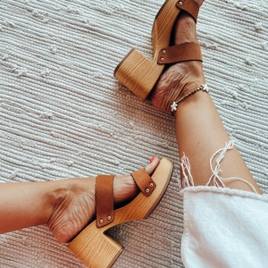 May include: A pair of brown wooden platform sandals with a brown leather strap. The sandals have a chunky heel and are worn on a person's feet.