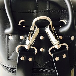 May include: Close-up of a black leather bag with silver hardware. The bag has a zipper closure and two straps with silver clasps.