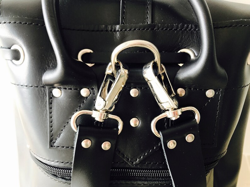 May include: Close-up of a black leather bag with silver hardware. The bag has a zipper closure and two straps with silver clasps.