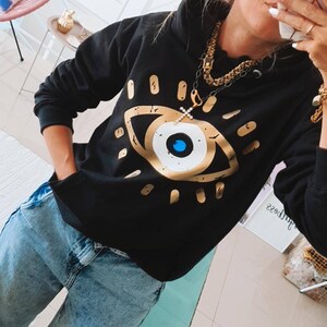 May include: Black hooded sweatshirt with a gold evil eye graphic print. The sweatshirt has a gold chain necklace with a cross pendant.