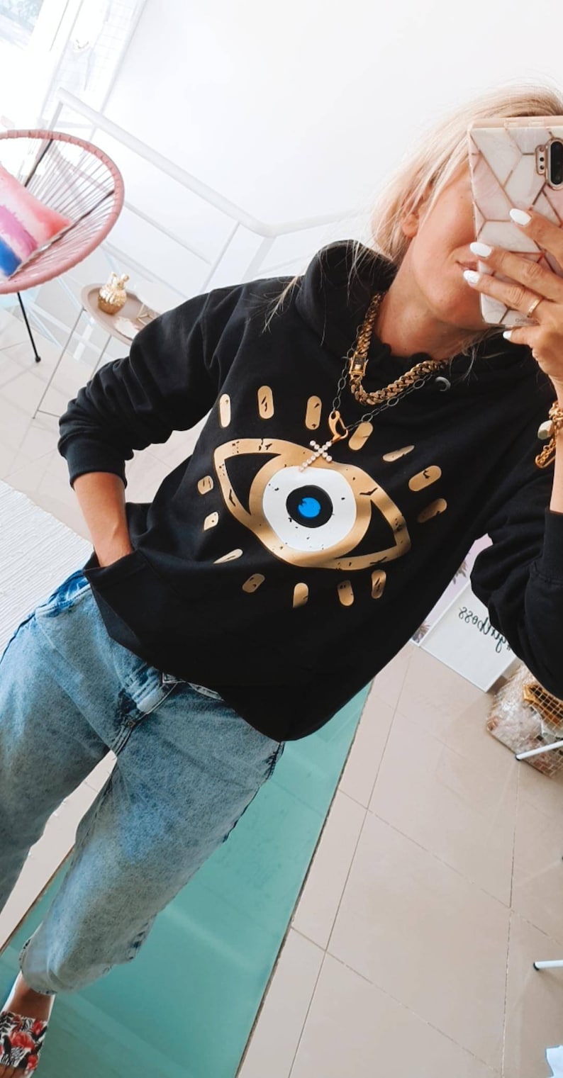May include: Black hooded sweatshirt with a gold evil eye graphic print. The sweatshirt has a gold chain necklace with a cross pendant.