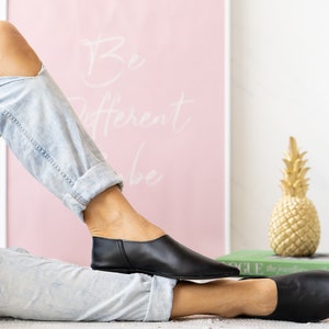 May include: A pair of black leather slip-on shoes with pointed toes. The shoes are worn by a person wearing ripped denim jeans. The shoes are on a light-colored surface. A gold pineapple figurine is in the background.