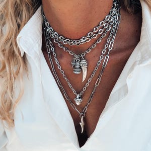 May include: A multi-layered silver necklace with different chain designs. The necklace includes charms such as a tooth, a small bell-shaped pendant, and a lightning bolt. The jewellery is worn over a white button-down shirt.