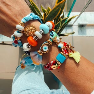 Può includere: Un set di braccialetti colorati con perline, ciondoli e tessuto. I braccialetti presentano un ciondolo con l'occhio blu malocchio, un ciondolo con una moneta d'oro e un fiocco giallo.