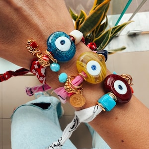 Puede incluir: Una pila de pulseras coloridas con dijes de ojo turco. Las pulseras están hechas de tela, cuentas y metal. Los dijes son azules, amarillos y rojos.