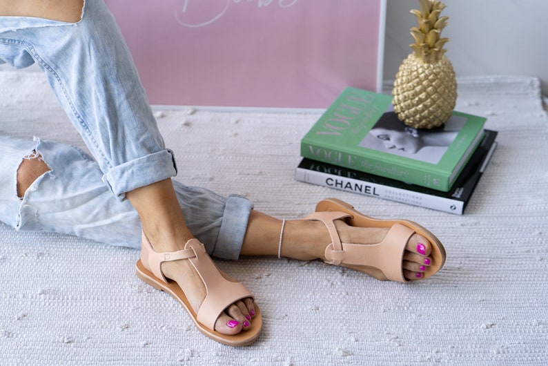 May include: A pair of light brown leather sandals with a t-strap design. The sandals have a flat sole and are worn by a person sitting on a white rug.