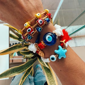 Könnte beinhalten: Ein Stapel bunter Armbänder mit bösen Augen-Anhängern und einem Seestern-Anhänger. Die Armbänder befinden sich am Handgelenk einer Person.