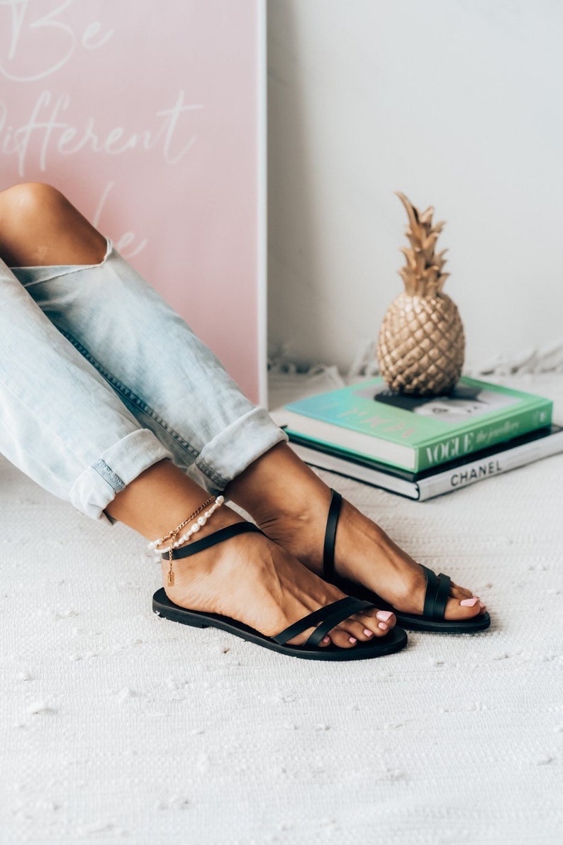 Puede incluir: Un par de sandalias de cuero negro con un dise&ntilde;o de tiras cruzadas. Las sandalias las lleva una persona con una cadena de oro y un tobillo de perlas. Las sandalias est&aacute;n sobre una superficie blanca y esponjosa.