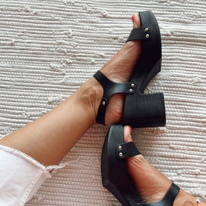 May include: Black platform sandals with silver studs and a thick wooden heel. The sandals have a strap across the foot and an ankle strap.