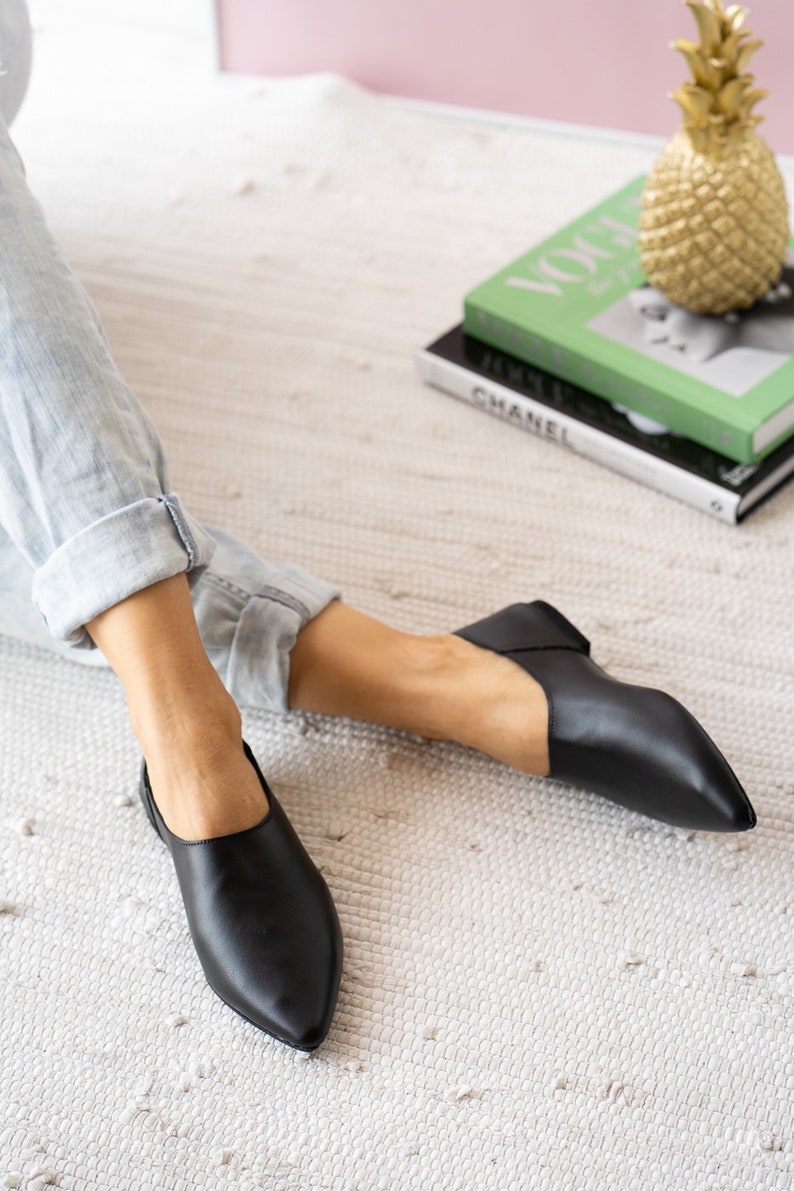 May include: A pair of black leather slip-on shoes with pointed toes. The shoes are worn by a person sitting on a white rug.