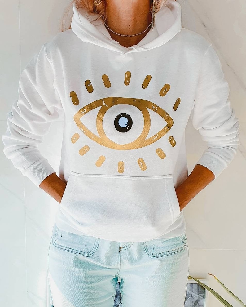 May include: White hooded sweatshirt with a gold evil eye graphic design. The eye has a blue pupil and is surrounded by gold rays.