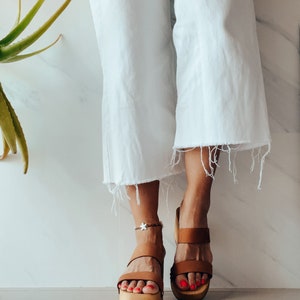 May include: Brown leather platform sandals with a white star ankle bracelet. The sandals have two straps across the foot and a thick platform sole.