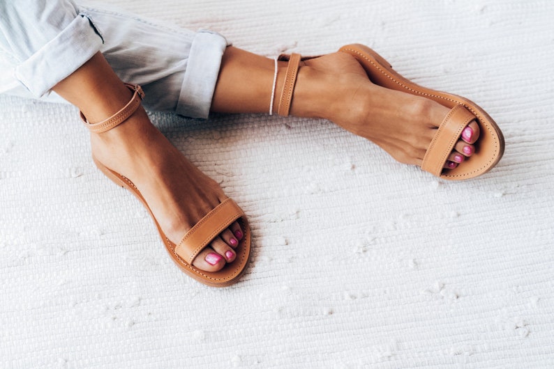 May include: A pair of brown leather sandals with a simple design. The sandals have a thin strap that goes around the ankle and a single strap across the top of the foot. The sandals are worn on a person's feet.