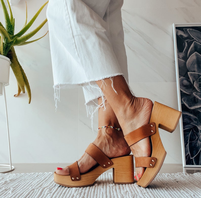 May include: A pair of brown leather platform clogs with wooden heels. The clogs have a strap across the foot and an ankle strap. The model is wearing white pants and is standing on a white surface.