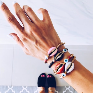 May include: A person's wrist adorned with several bracelets featuring gold-toned seashells and evil eye charms. The bracelets are layered and include pink, red, black, and white seashells.
