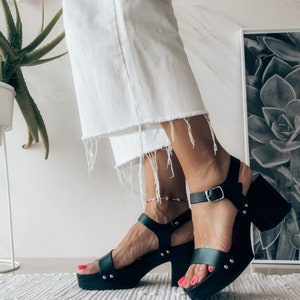 May include: Black leather platform sandals with a buckle closure and a chunky heel. The sandals have a platform sole and are worn on a person's feet.