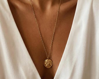 May include: A gold-colored necklace with a pendant featuring a detailed relief design. The pendant hangs from a delicate chain. The necklace is worn against a white top.
