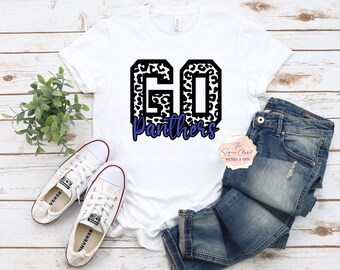 May include: White t-shirt with a black and white leopard print "GO Panthers" design. The shirt is displayed on a wooden surface with a pair of white sneakers and blue jeans.